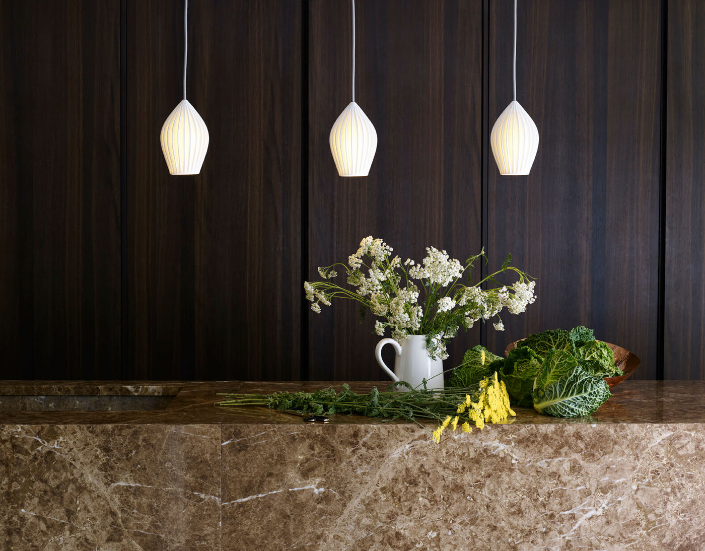 Modern kitchen with brown marble counter, dark wood wall, white pendant lights, and fresh flowers and greens