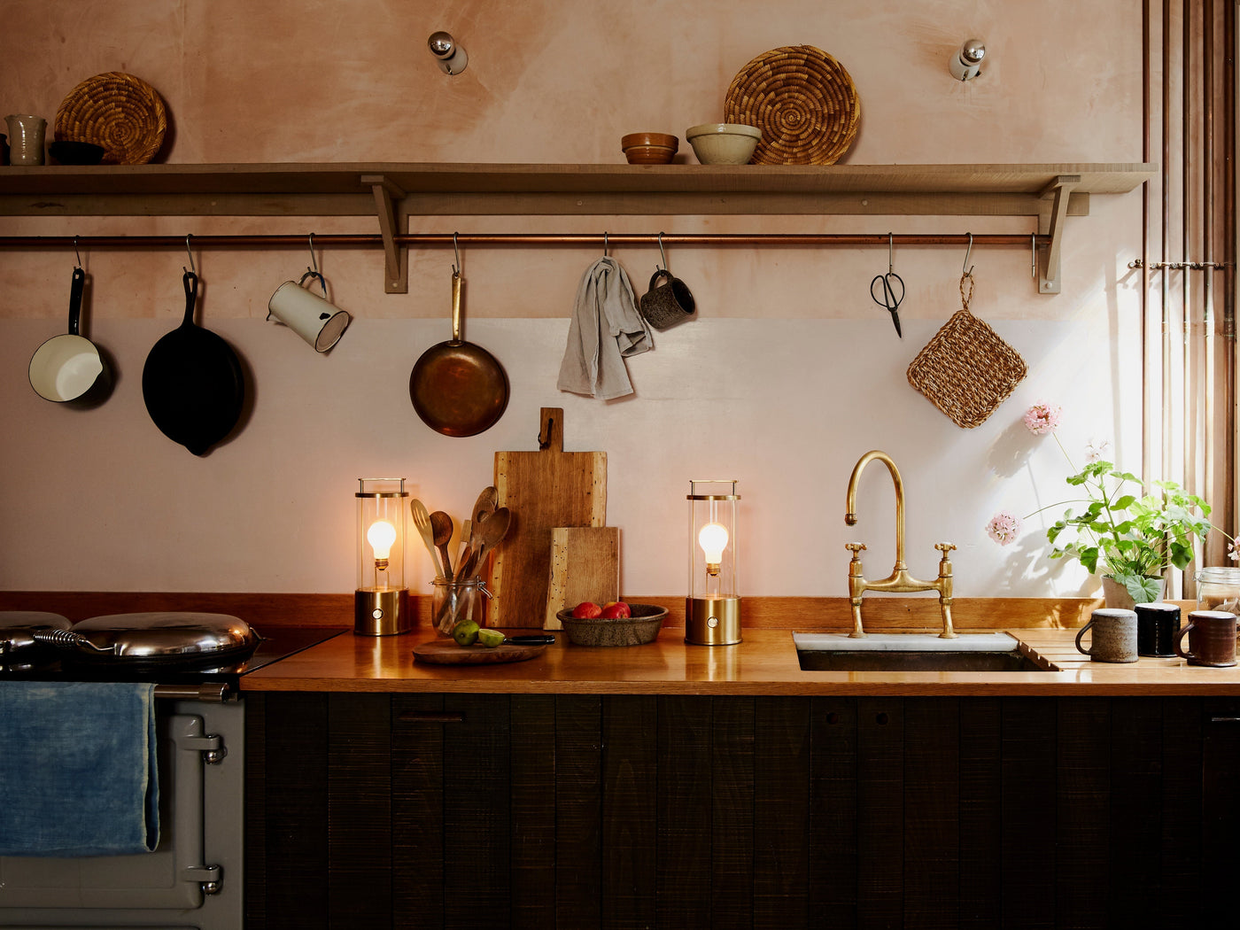 Cozy kitchen countertop with brass faucet, hanging pans, wooden utensils, soft lighting, and potted plants