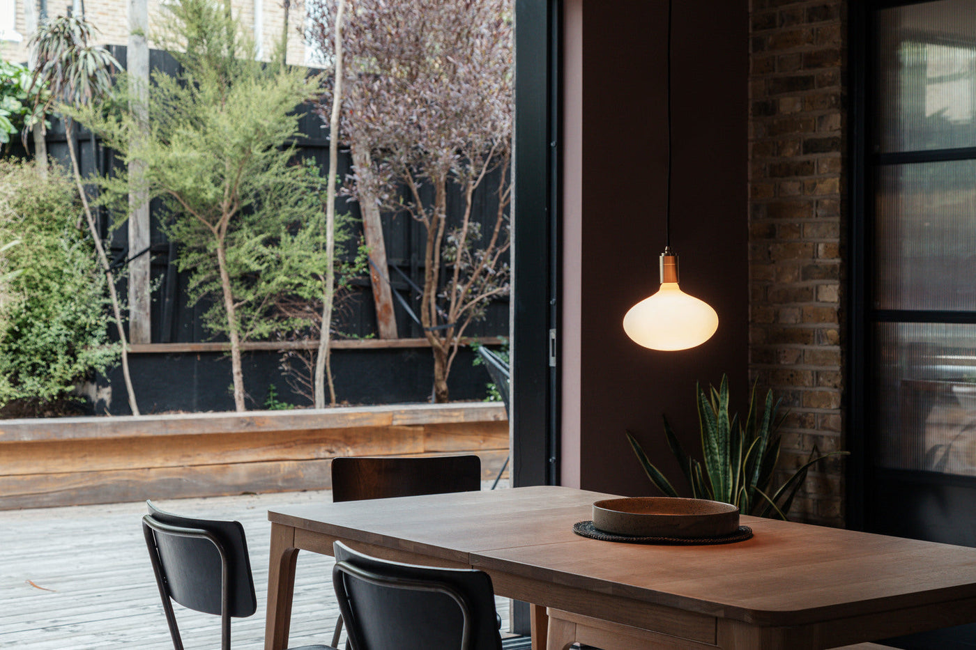 Modern dining area with wooden table, black chairs, hanging oval pendant light, and outdoor greenery