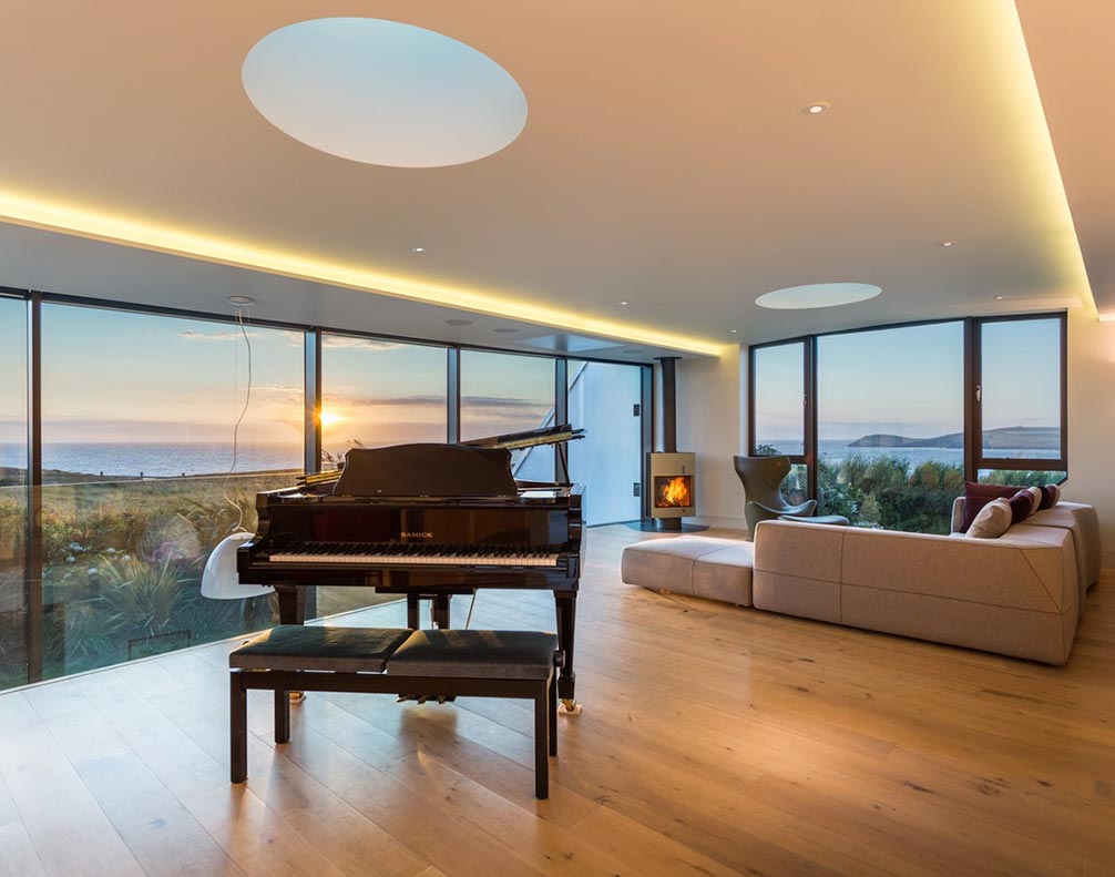 Modern living room with grand piano, curved beige sofa, fireplace, large windows overlooking ocean sunset