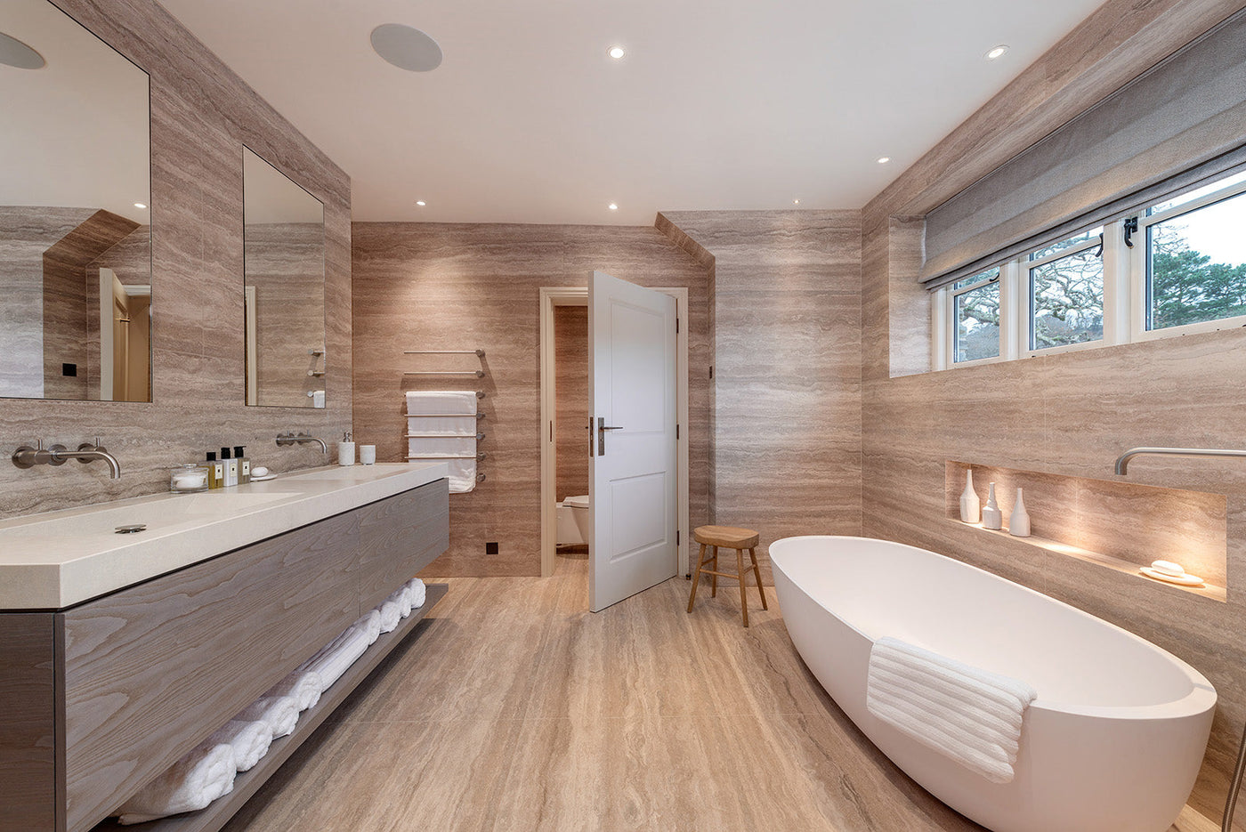 Modern beige stone bathroom with freestanding tub, double sinks, towel rack, and wooden stool