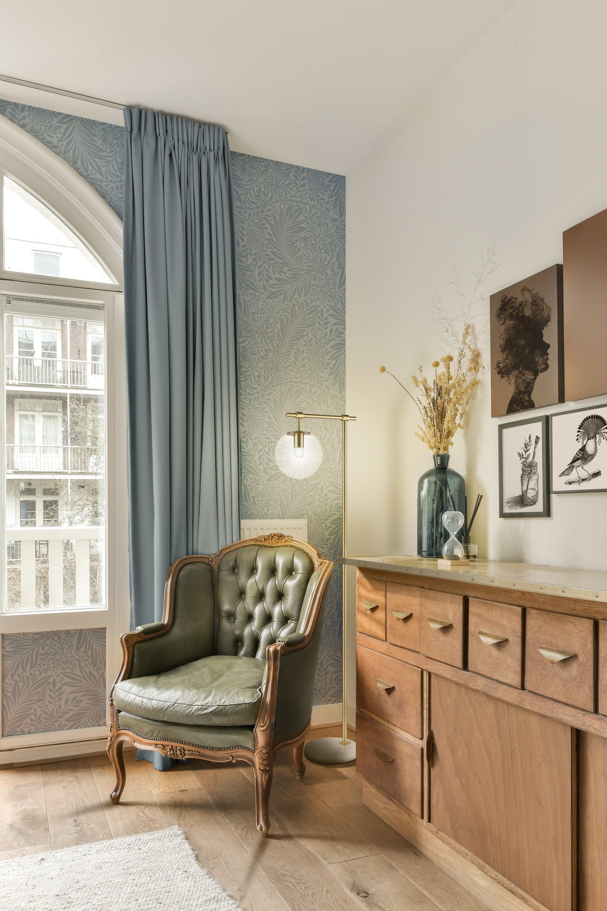 Cozy living room corner with vintage green tufted armchair, wooden sideboard, blue curtains, brass floor lamp, and botanical wall art