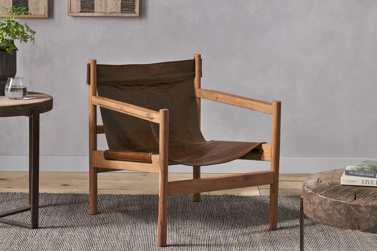 A tan Nkuku Dalibar Leather Lounger chair with a wooden frame, situated in a room with a grey wall and a wooden side table.