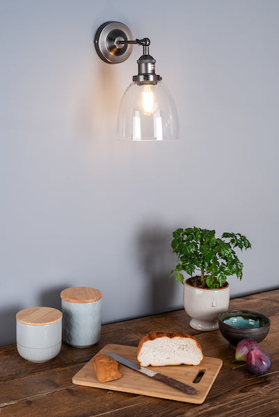 Antique chrome wall light with clear glass shade illuminating kitchen countertop with bread, jars, plant, and figs