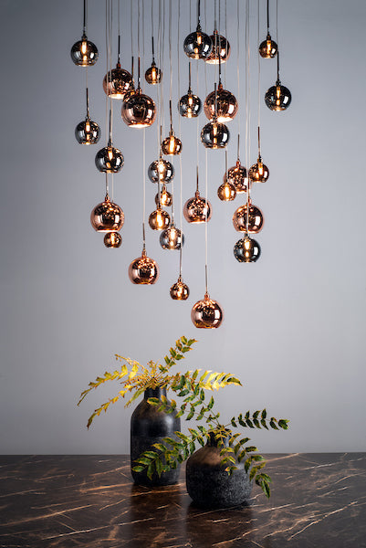 Modern cluster pendant light with 30 copper and bronze glass globes hanging over a marble table with two black vases and green foliage