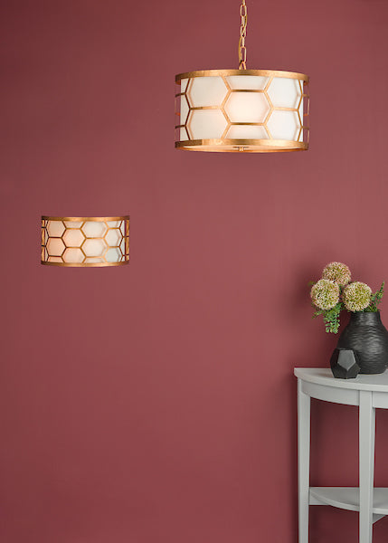 Gold hexagon-patterned 3-light pendant with ivory shade against a burgundy wall above a white side table with black vase and flowers
