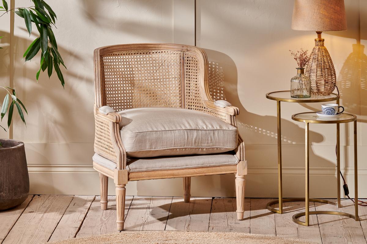 A boho-inspired Nkuku Kaziria Cane & Linen Armchair in natural color, featuring a cane backrest and wooden legs, positioned in a room with a side table and lamp.