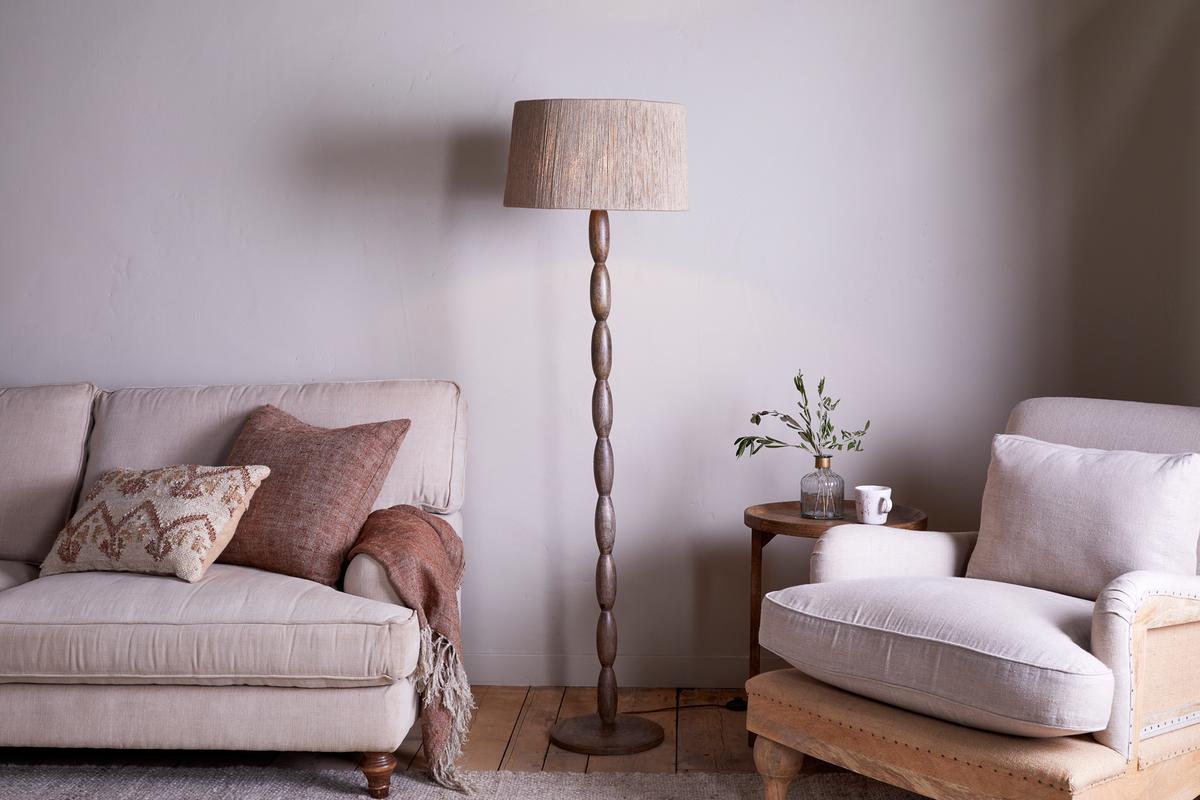 A sleek Nkuku Konali Mango Wood Floor Lamp with an ash stain finish, standing in a living room setting with a sofa and armchair.