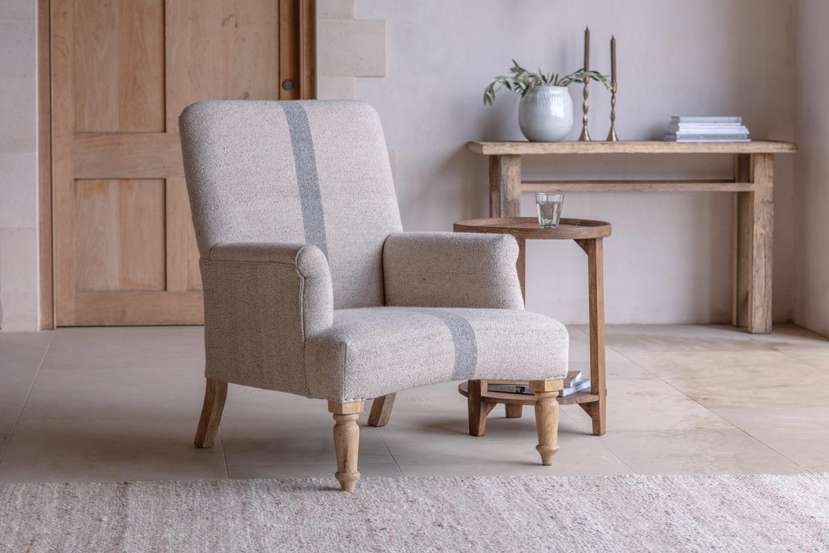 A beige Nkuku Kallu Wool & Cotton Occasional Chair with a wooden frame, placed in a room with a wooden table and a plant in the background.