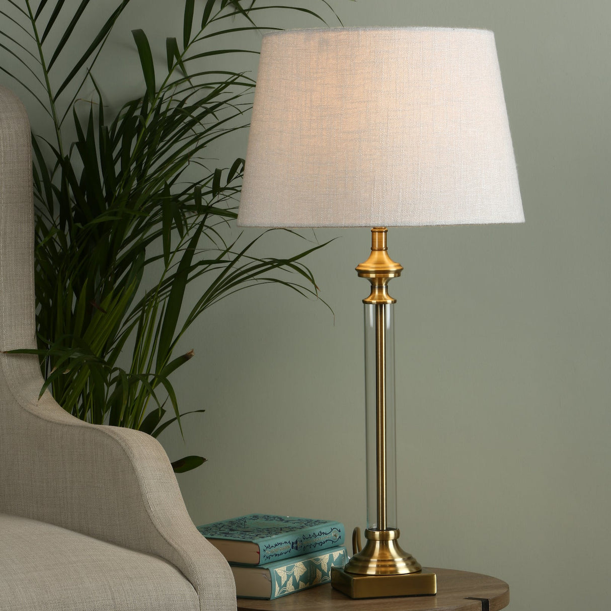 Brass table lamp with white fabric shade on wooden side table beside beige armchair and green plant