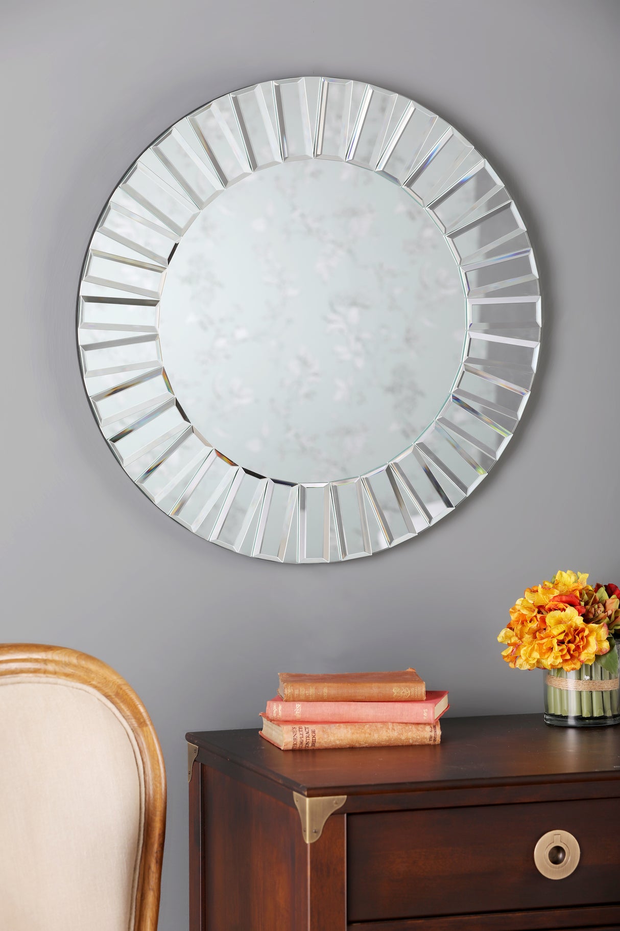Round decorative wall mirror with faceted frame above wooden side table with vintage books and orange flowers