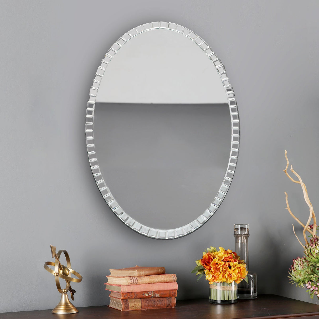 Oval decorative wall mirror with beveled frame above wooden table with vintage books, brass armillary sphere, and flower vases