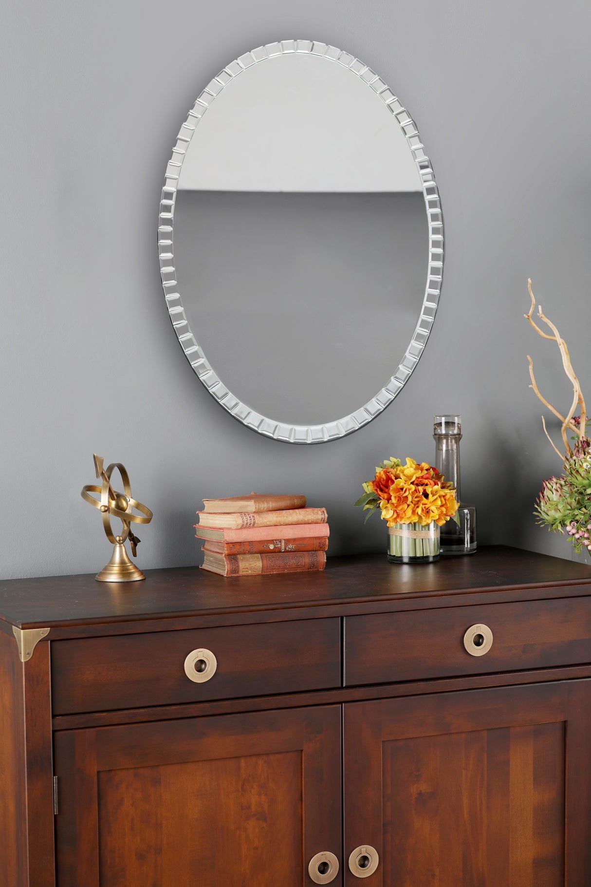 Oval decorative mirror above wooden cabinet with vintage books, brass armillary sphere, and flower arrangements
