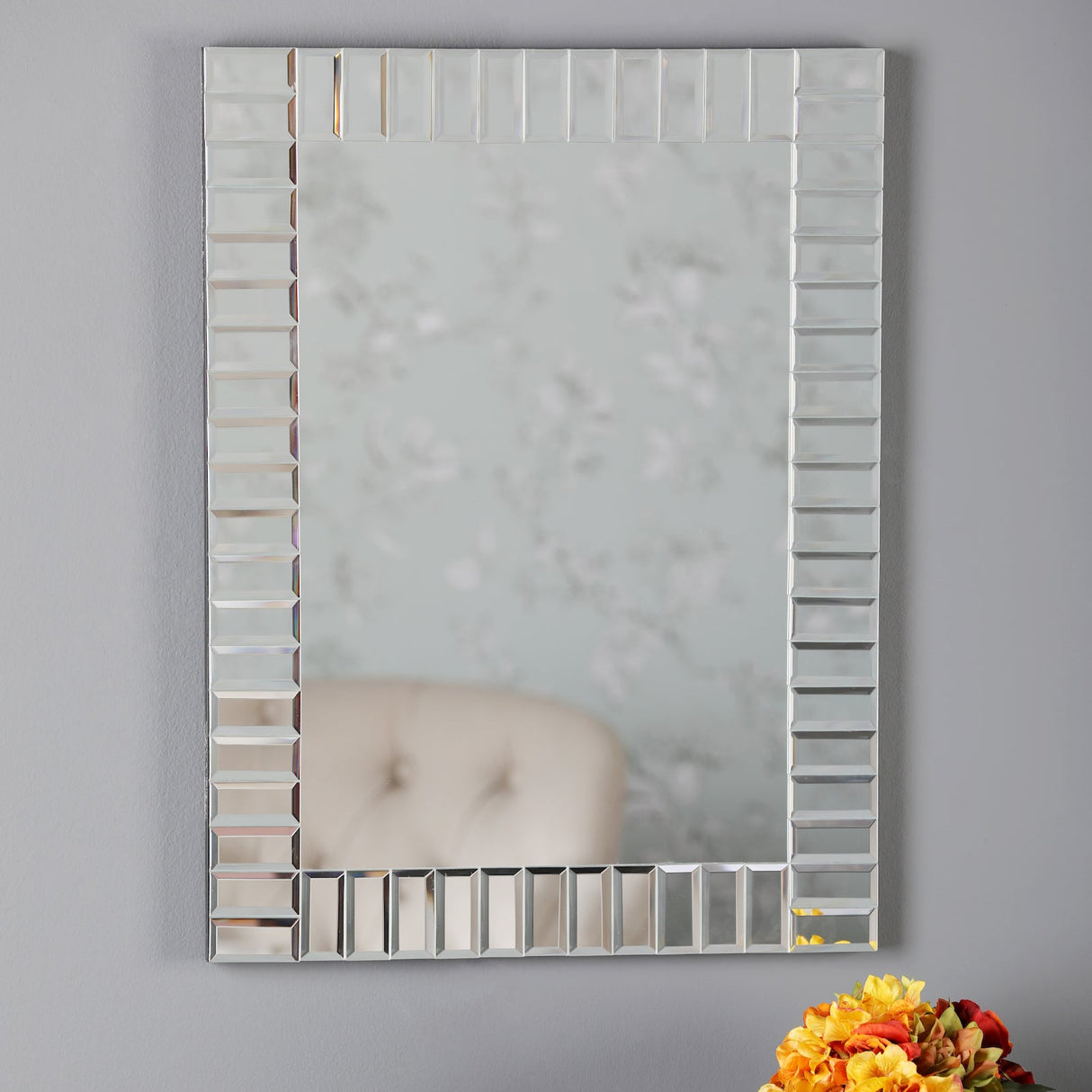 Decorative square wall mirror with beveled glass frame reflecting floral wallpaper and beige chair