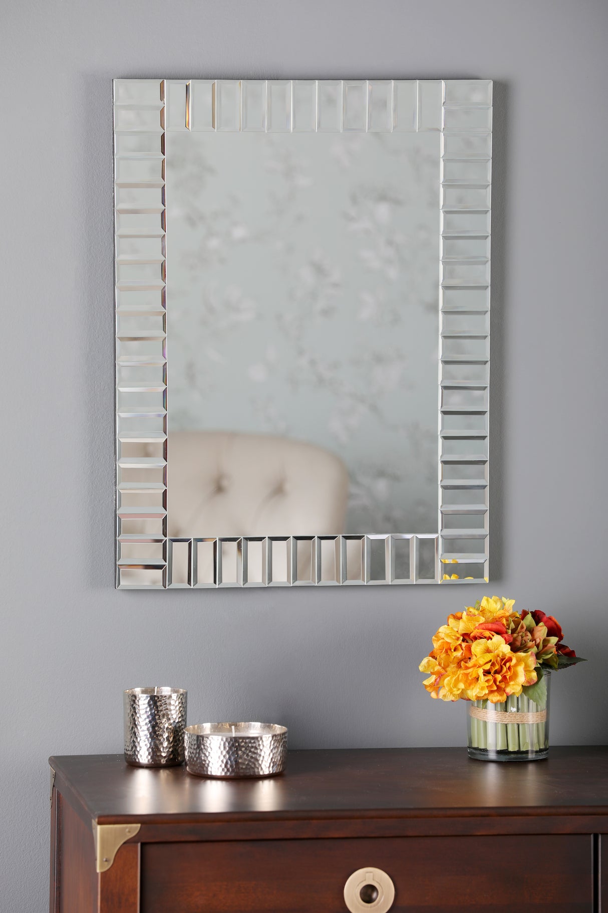Decorative rectangular wall mirror above dark wooden dresser with silver candle holders and orange flowers