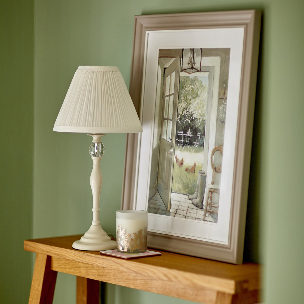 White table lamp with pleated shade and decorative candle on wood shelf with framed garden painting