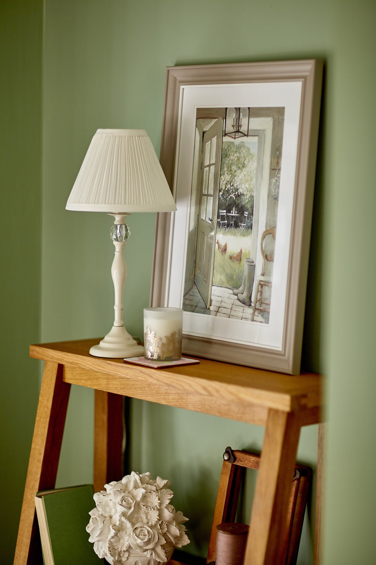 Wooden console table with white lamp, scented candle, and framed garden painting against green wall