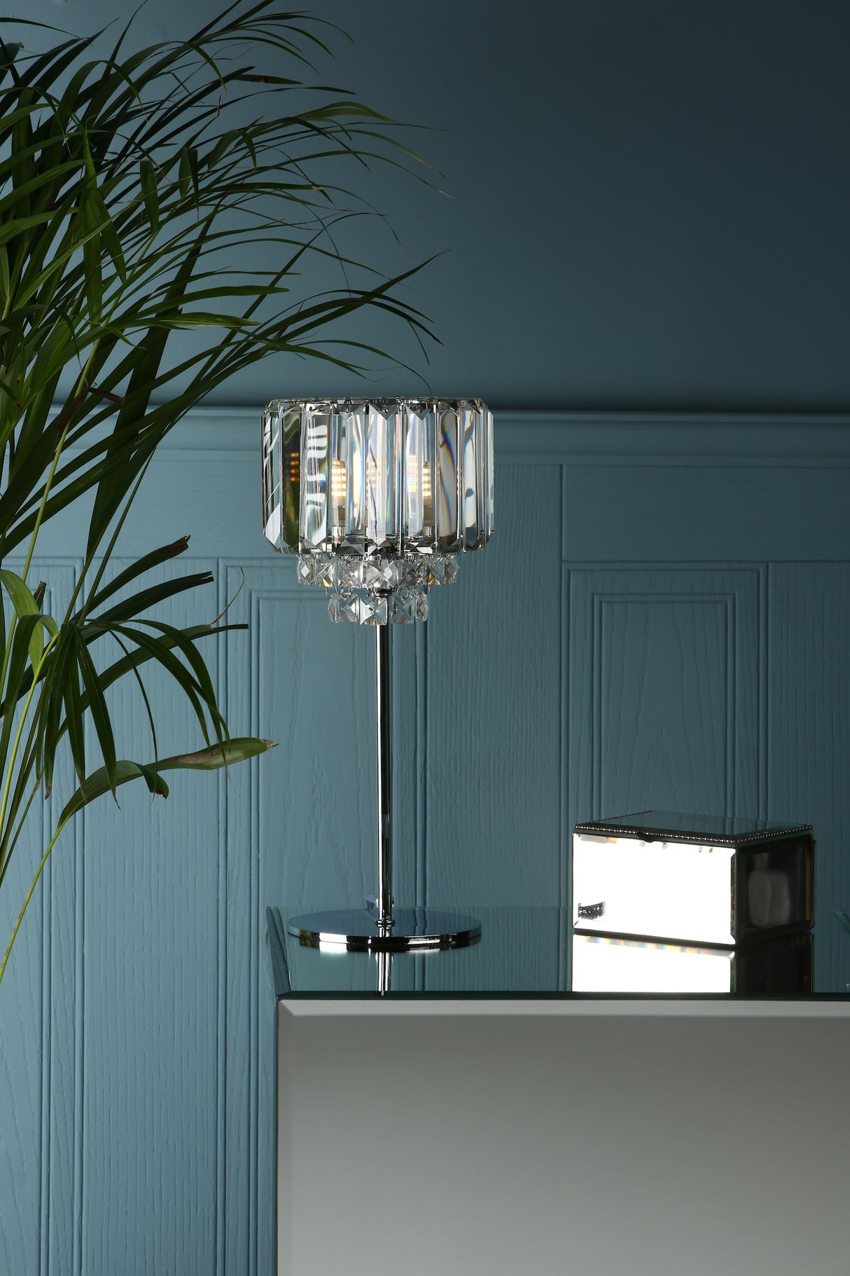 Modern crystal table lamp with metallic base on glass surface against blue paneled wall with green plant