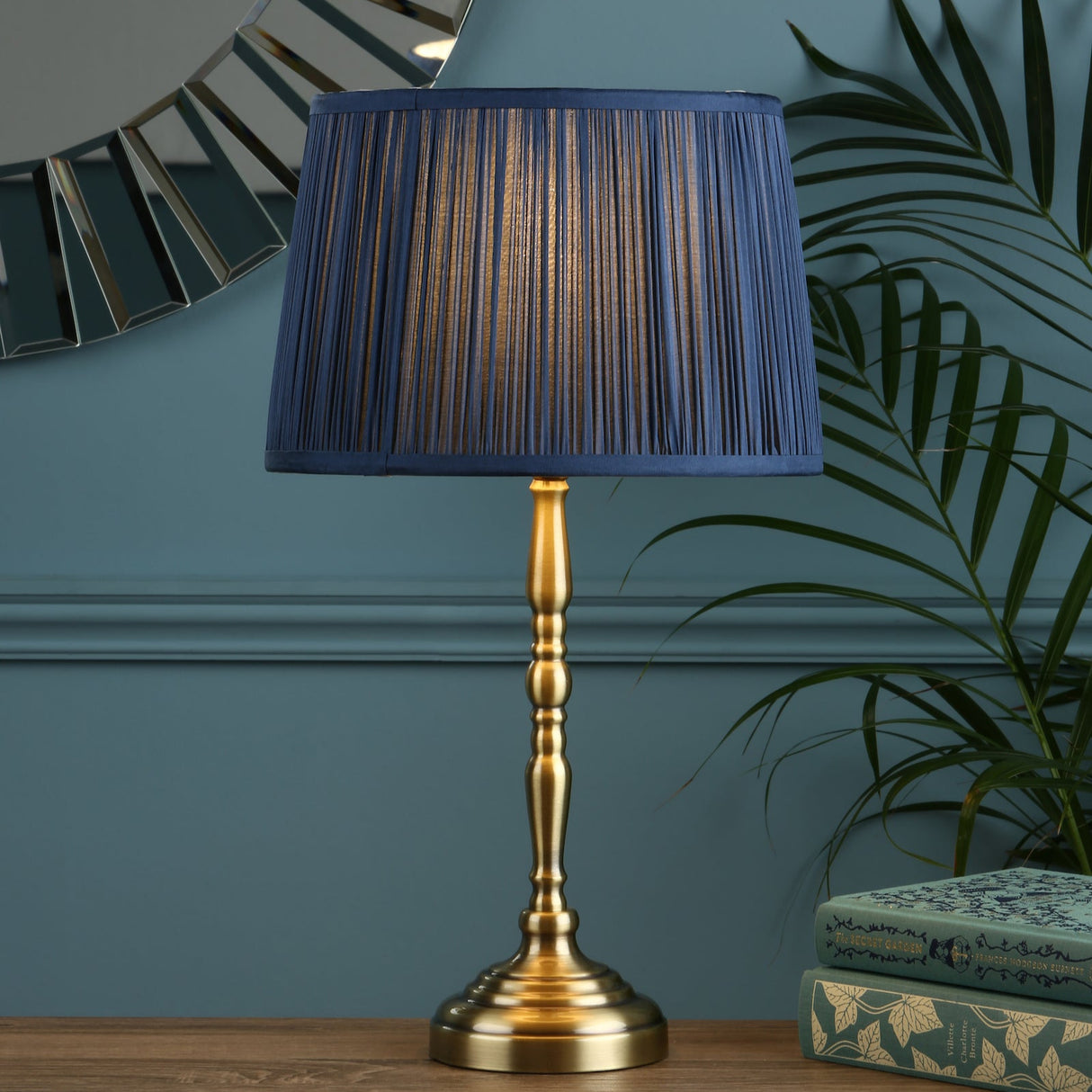 Brass table lamp with pleated navy blue shade on wooden surface, decorative books and green plant