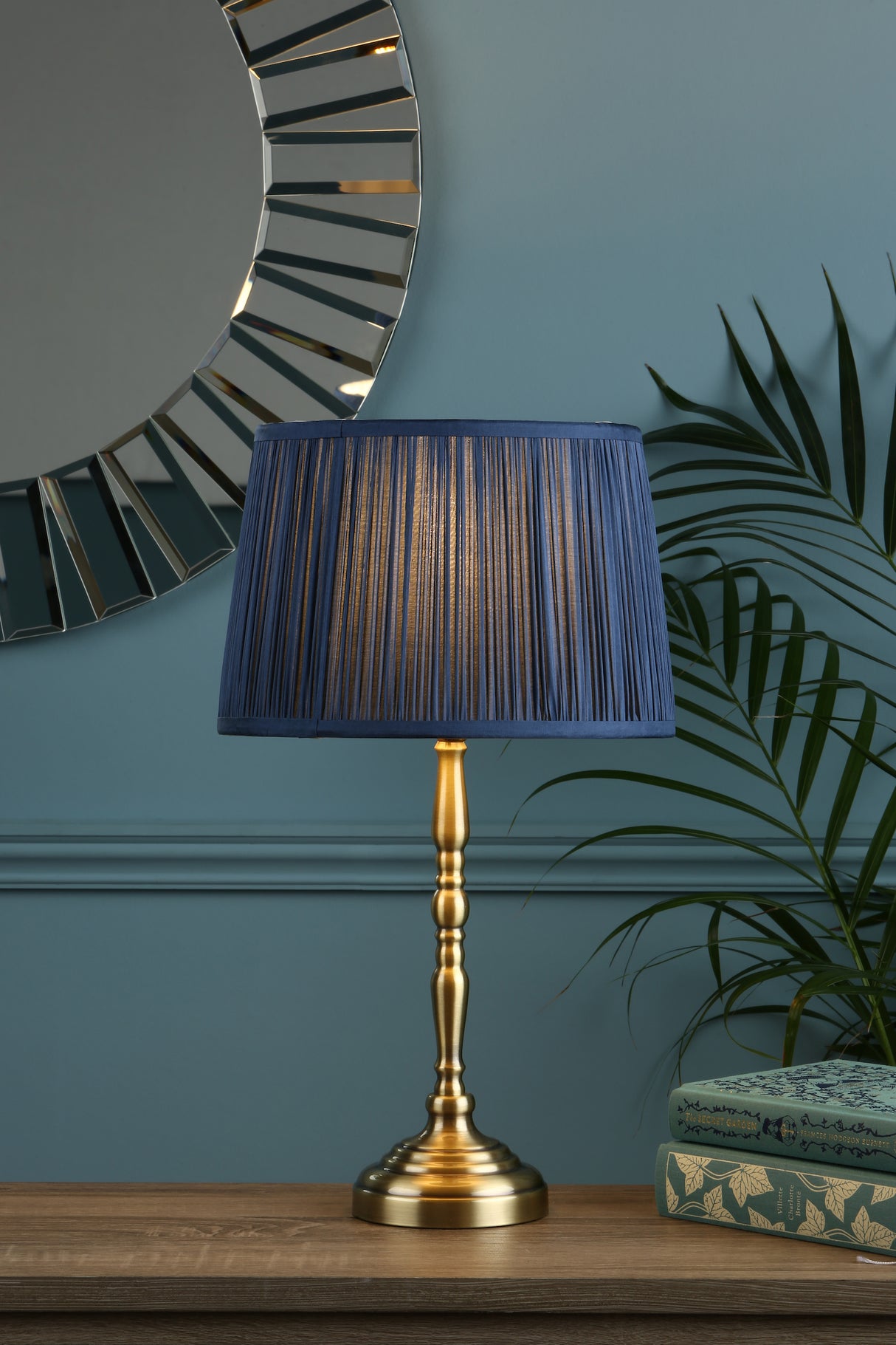 Brass table lamp with pleated blue shade on wooden surface, round decorative mirror, green wall and plant