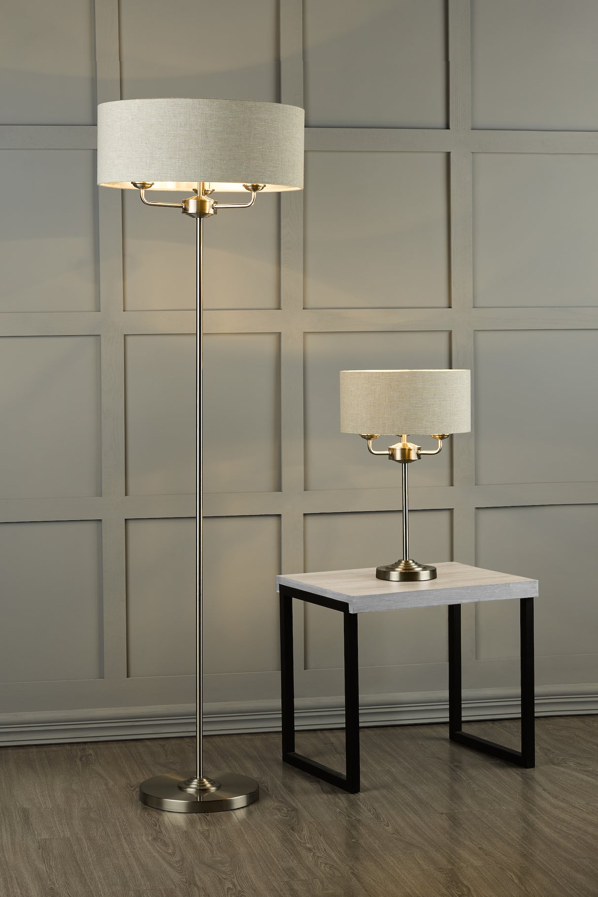 modern brass floor lamp and matching table lamp with beige shades in a room with gray paneled wall