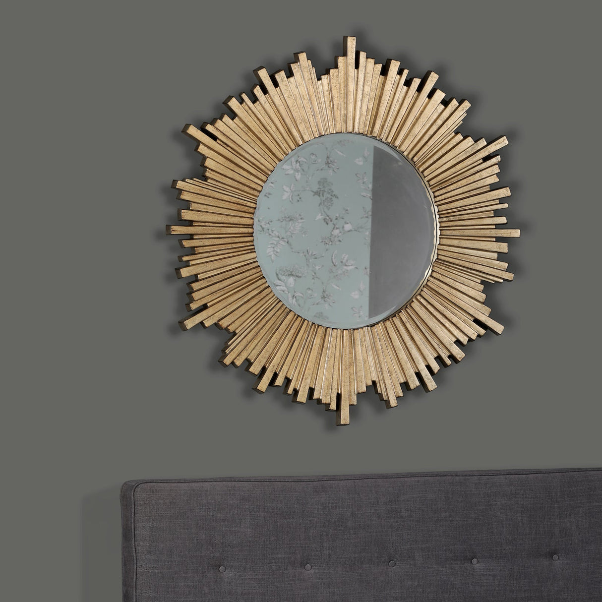 Round sunburst wall mirror with gold textured frame above dark gray upholstered headboard on gray wall