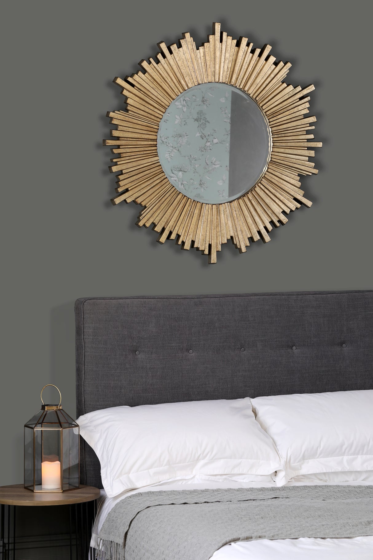 Modern bedroom with dark grey upholstered headboard, white pillows, grey blanket, sunburst gold mirror, and candle lantern
