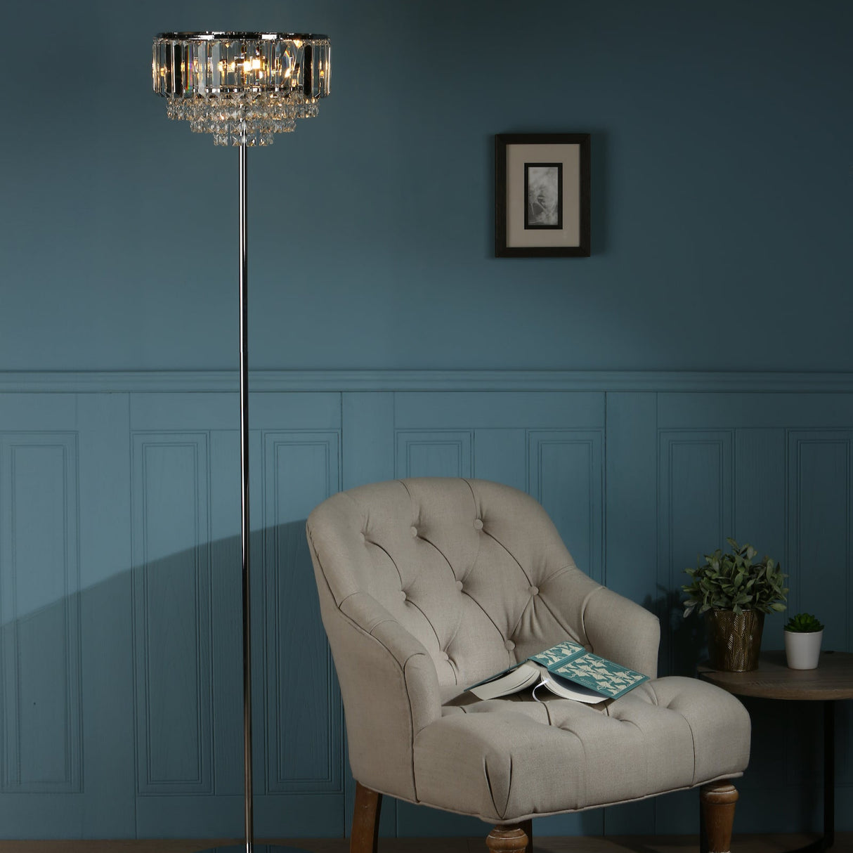 Beige tufted armchair with open book under crystal floor lamp in a blue paneled room