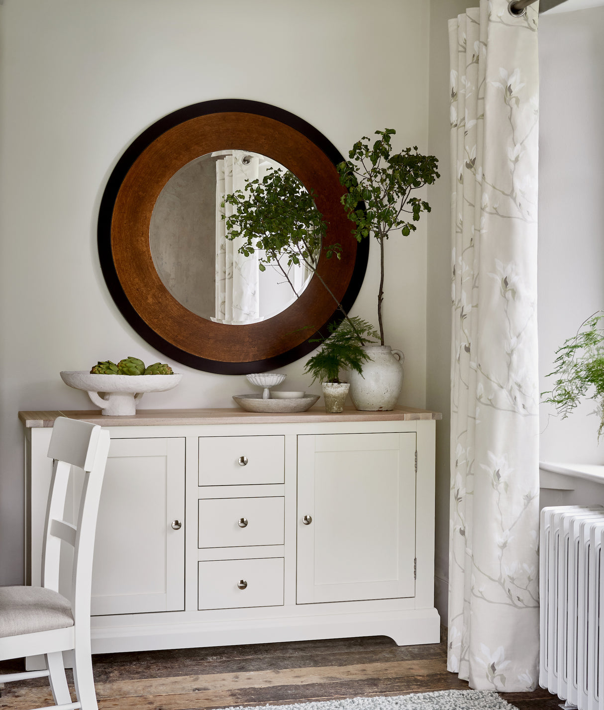 White sideboard with round wooden-framed mirror, plants in ceramic vases, and artichokes on platter