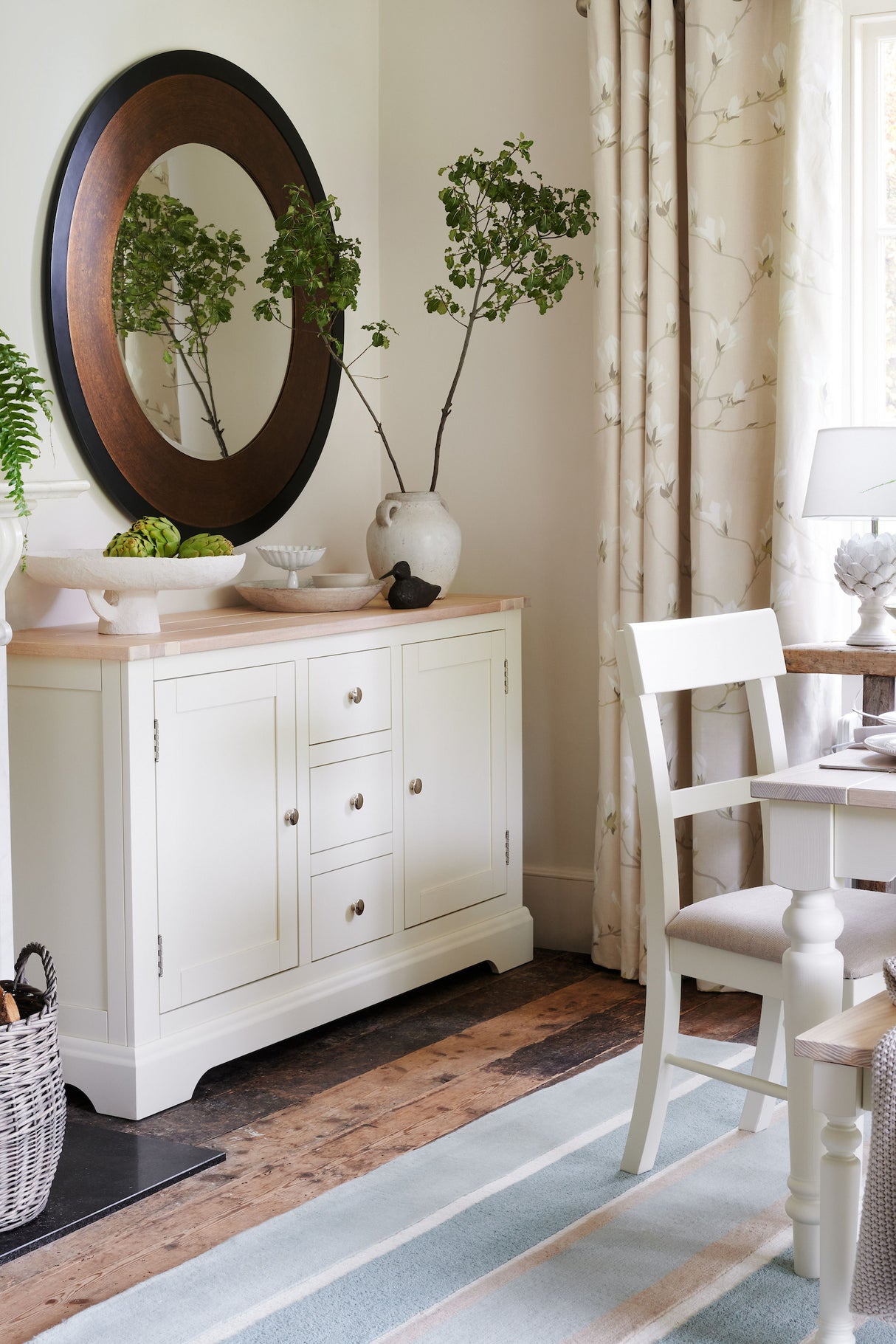 Cozy dining room with white wooden sideboard, round mirror, leafy branches in vase, and white chair