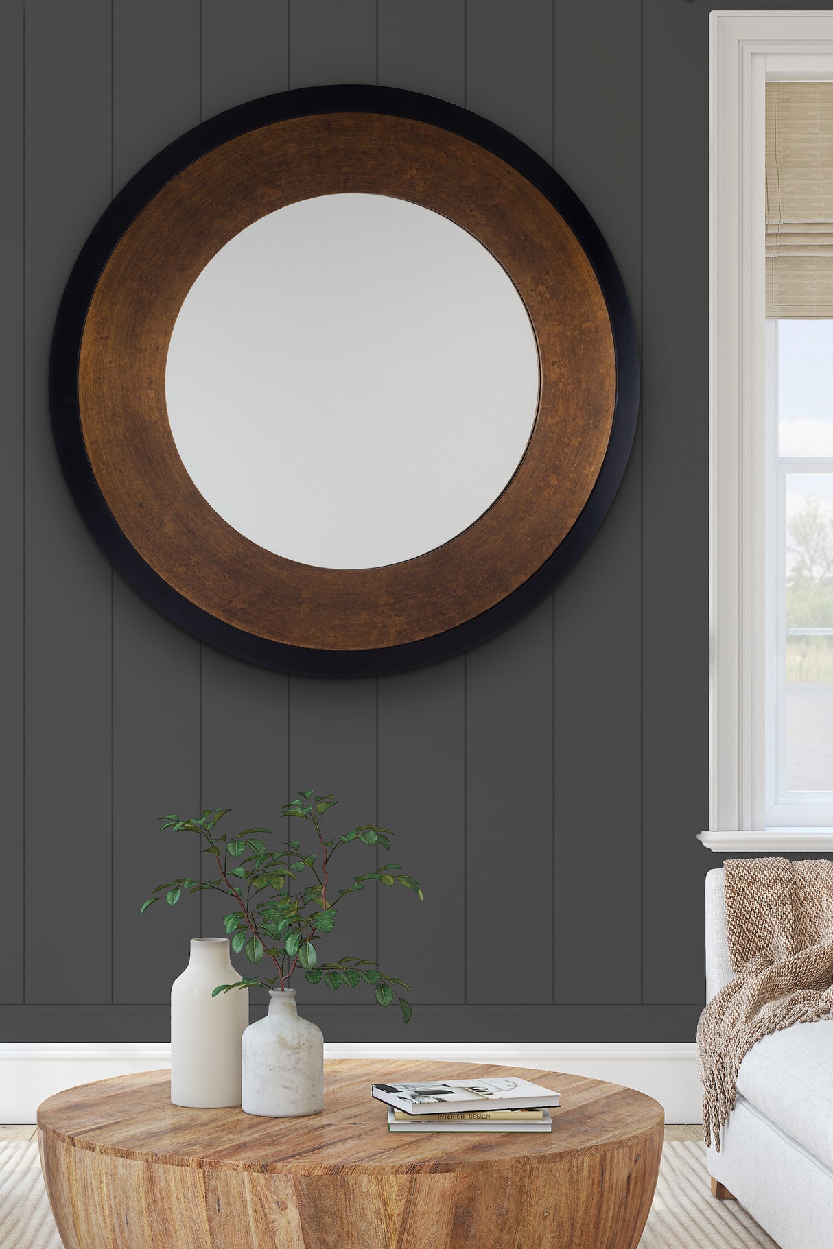 Round wall mirror with wooden frame on dark gray paneled wall above wooden coffee table with vases and books