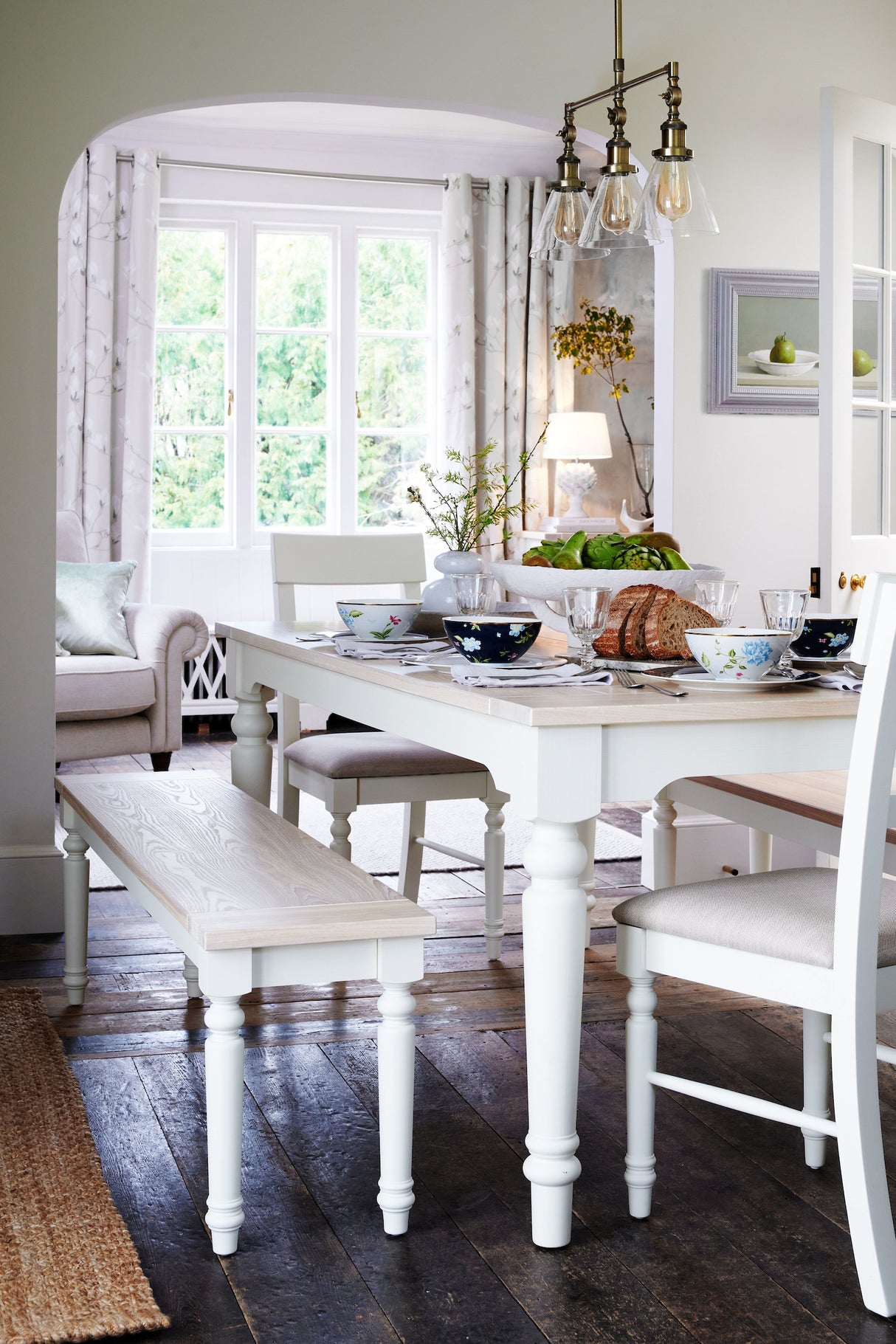 Bright dining room with white wood table, bench, chairs, floral bowls, bread, and vintage pendant lights