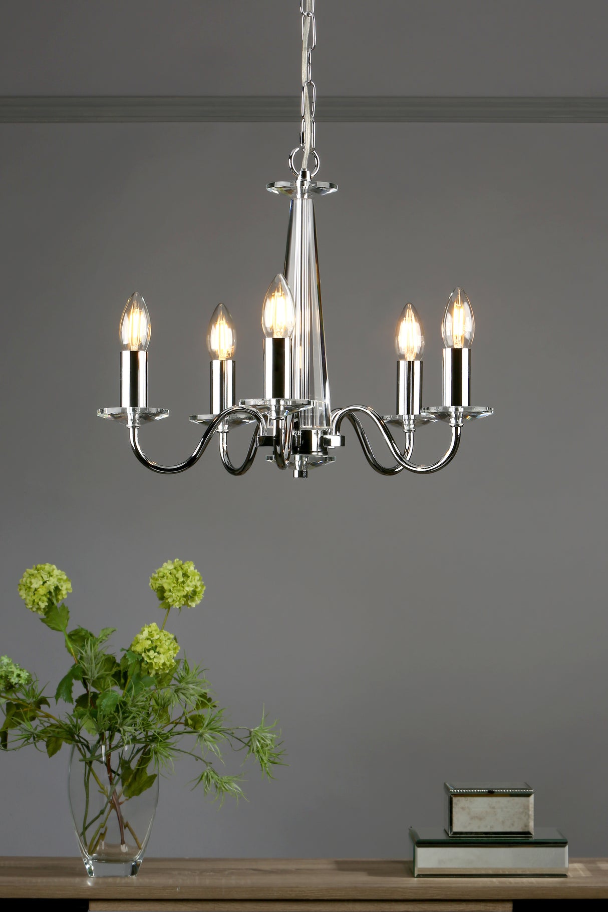 Modern silver chandelier with five lit bulbs above a wooden table with green floral arrangement and mirrored boxes