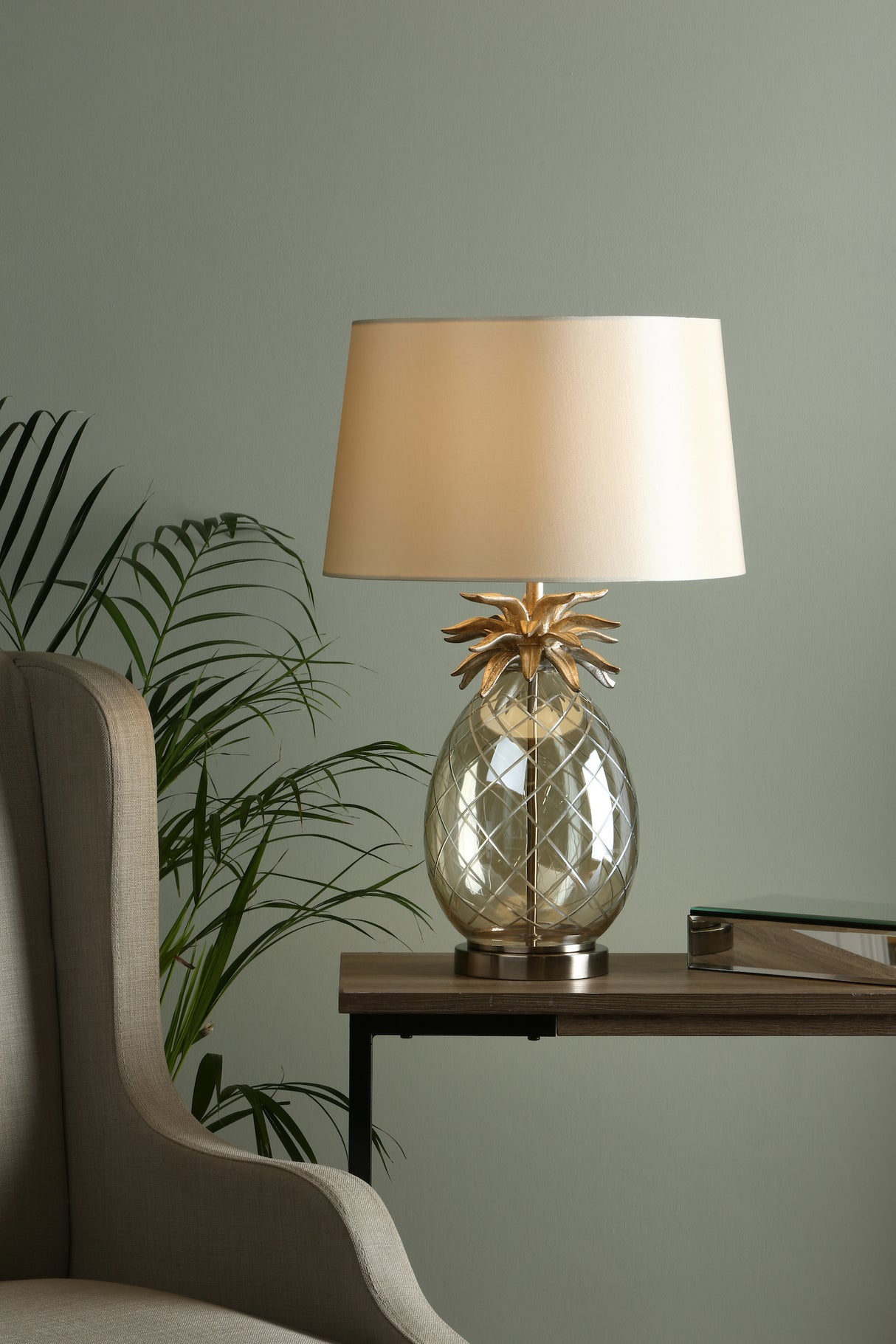 Glass table lamp with golden pineapple design and beige shade on wooden side table