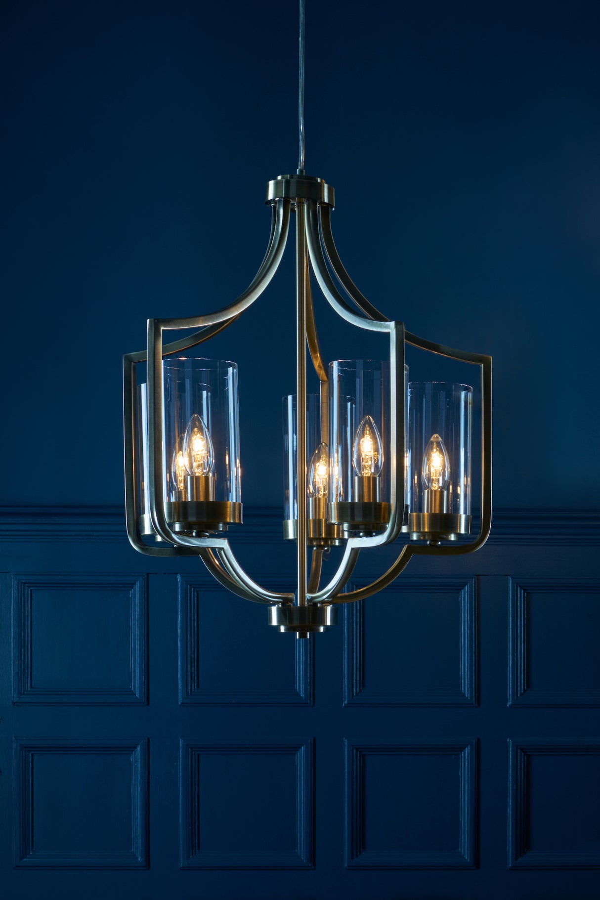 Modern brass chandelier with five candle-style bulbs encased in glass, against dark blue paneled wall
