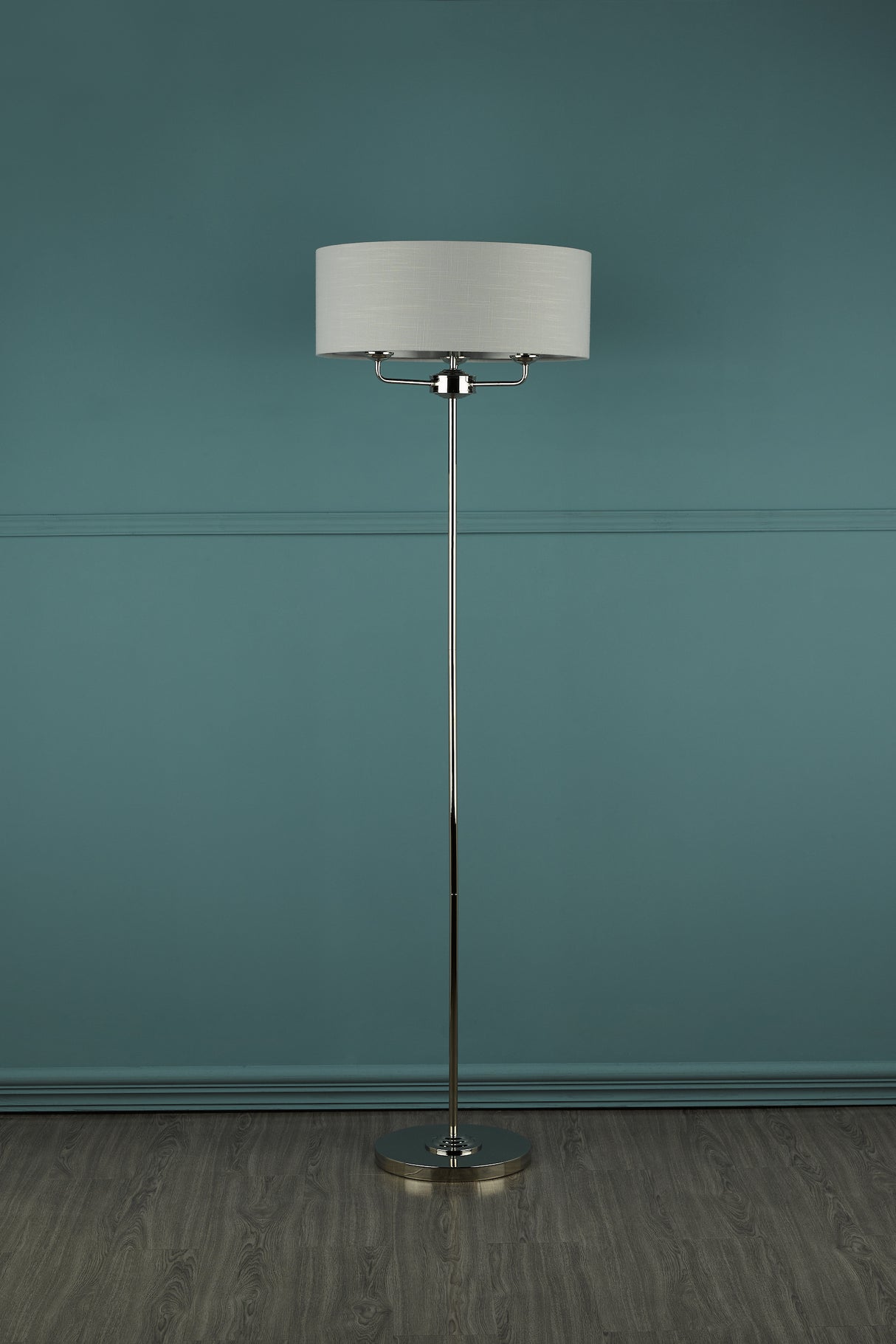 Modern floor lamp with white fabric shade and chrome stand on wood floor against teal wall