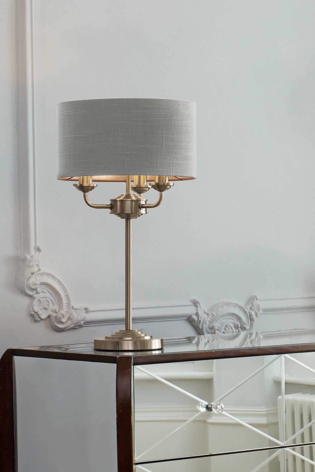 Modern brushed brass table lamp with gray fabric drum shade on mirrored cabinet in elegant room
