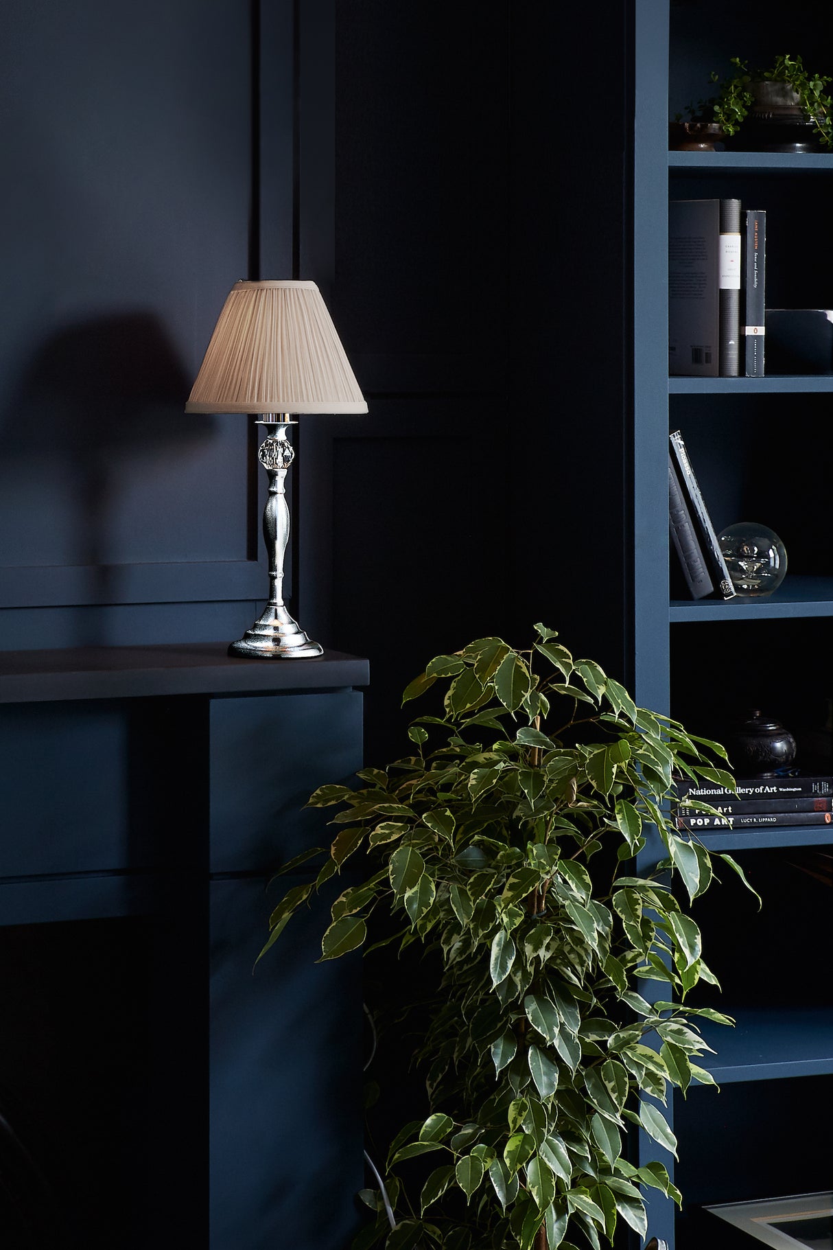 Table lamp with beige pleated shade on dark blue shelf next to green leafy plant and bookshelf