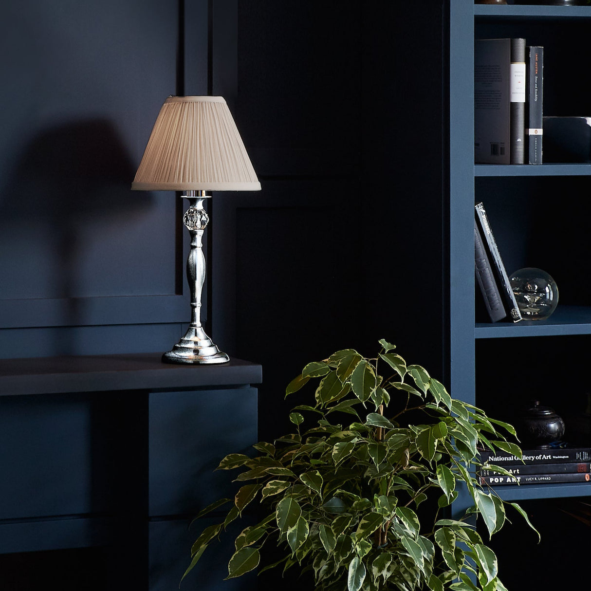 Silver table lamp with beige pleated shade on dark shelf near green leafy plant and blue bookshelf with books