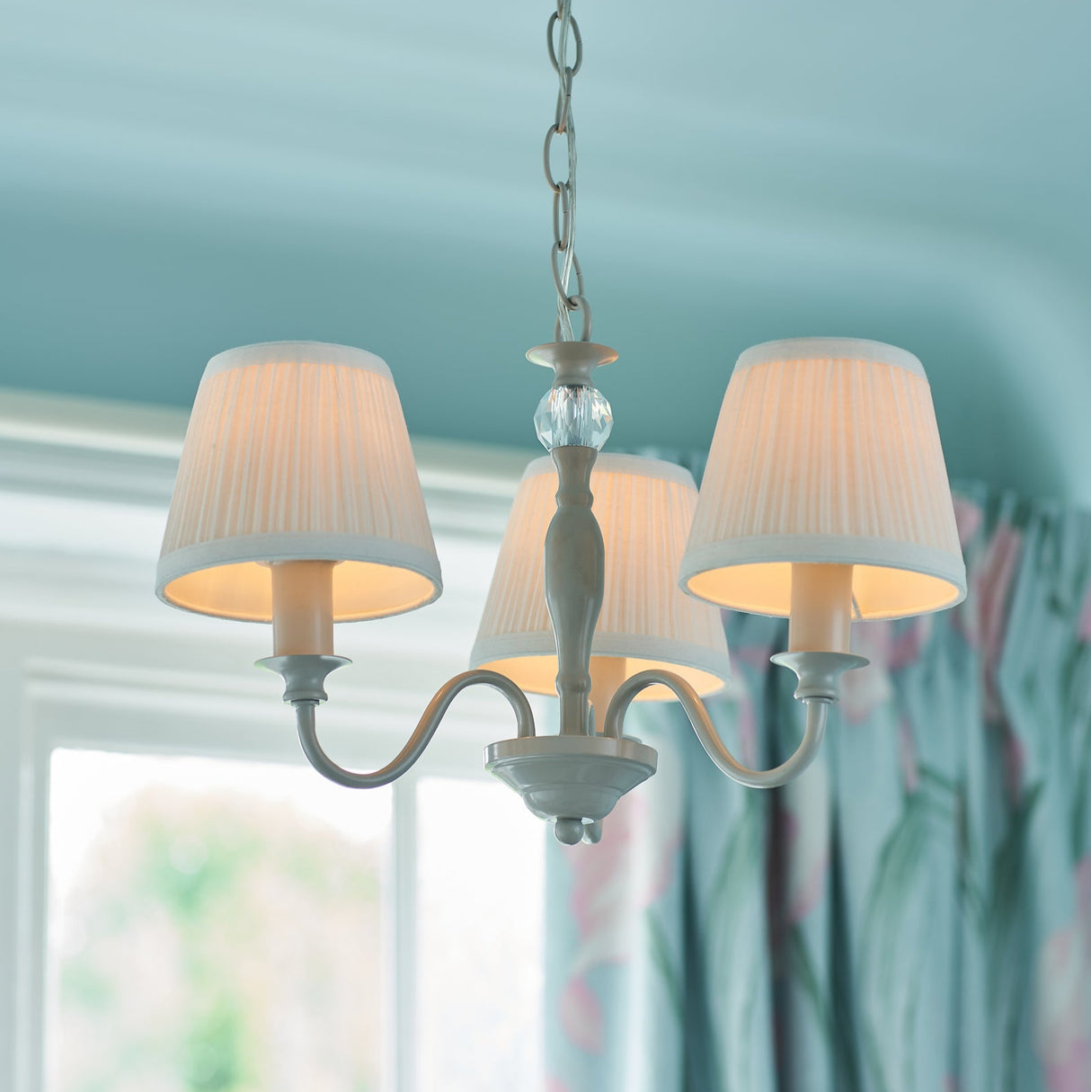 Elegant white chandelier with three lampshades hanging in a room with floral curtains and blue ceiling