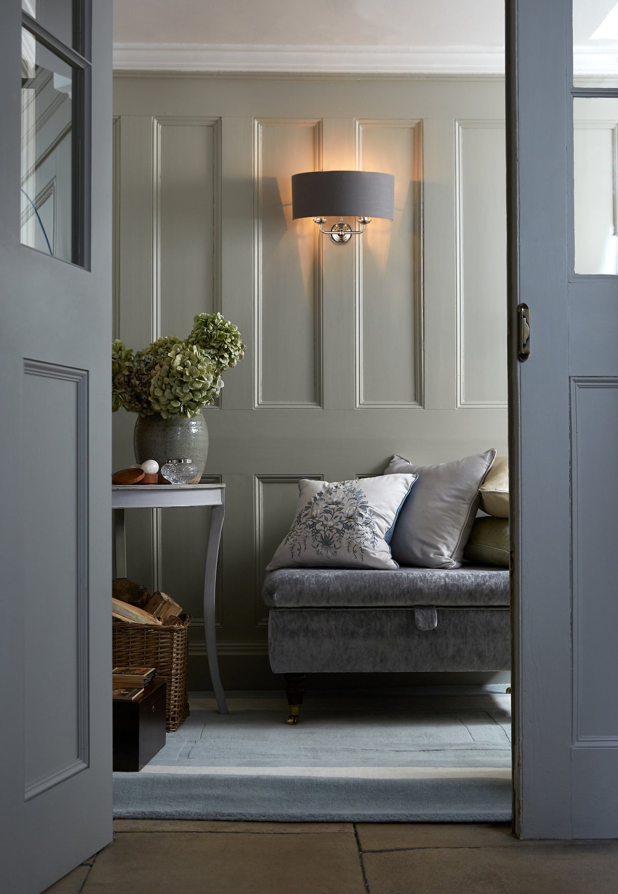 Cozy sitting area with velvet bench, decorative pillows, gray wall sconce, and vase of dried flowers