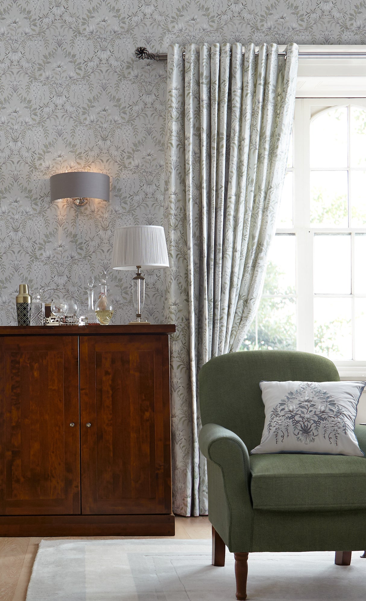 Cozy living room corner with green upholstered armchair, decorative pillow, wooden cabinet, patterned curtains, and floral wallpaper