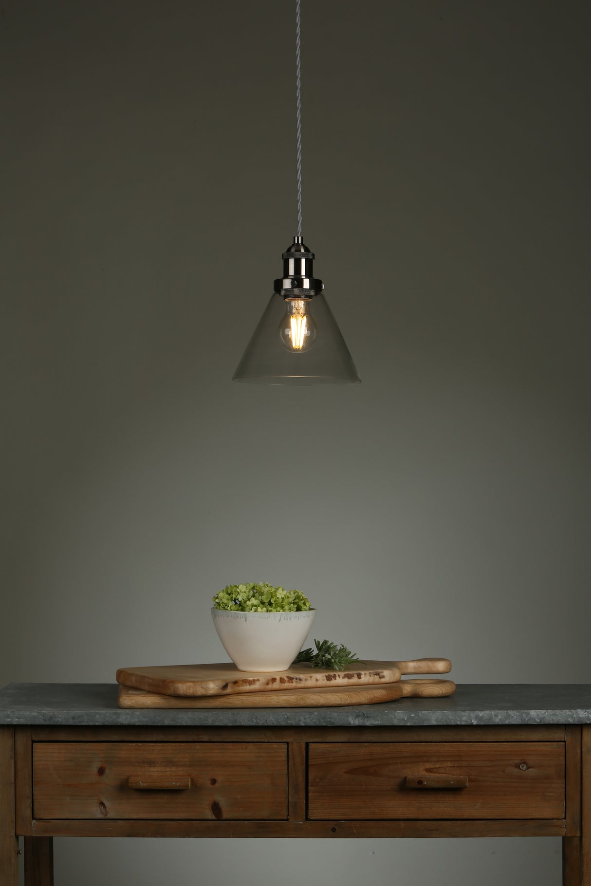 Minimalist kitchen scene with wooden table, two cutting boards, white bowl with greens, and hanging pendant light