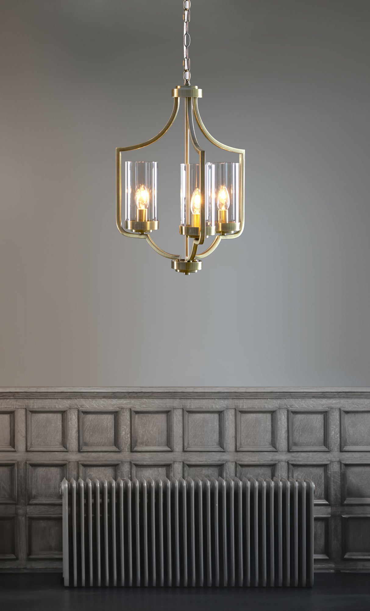 Brass chandelier with four lit bulbs and glass shades hanging above a vintage radiator and paneled wall