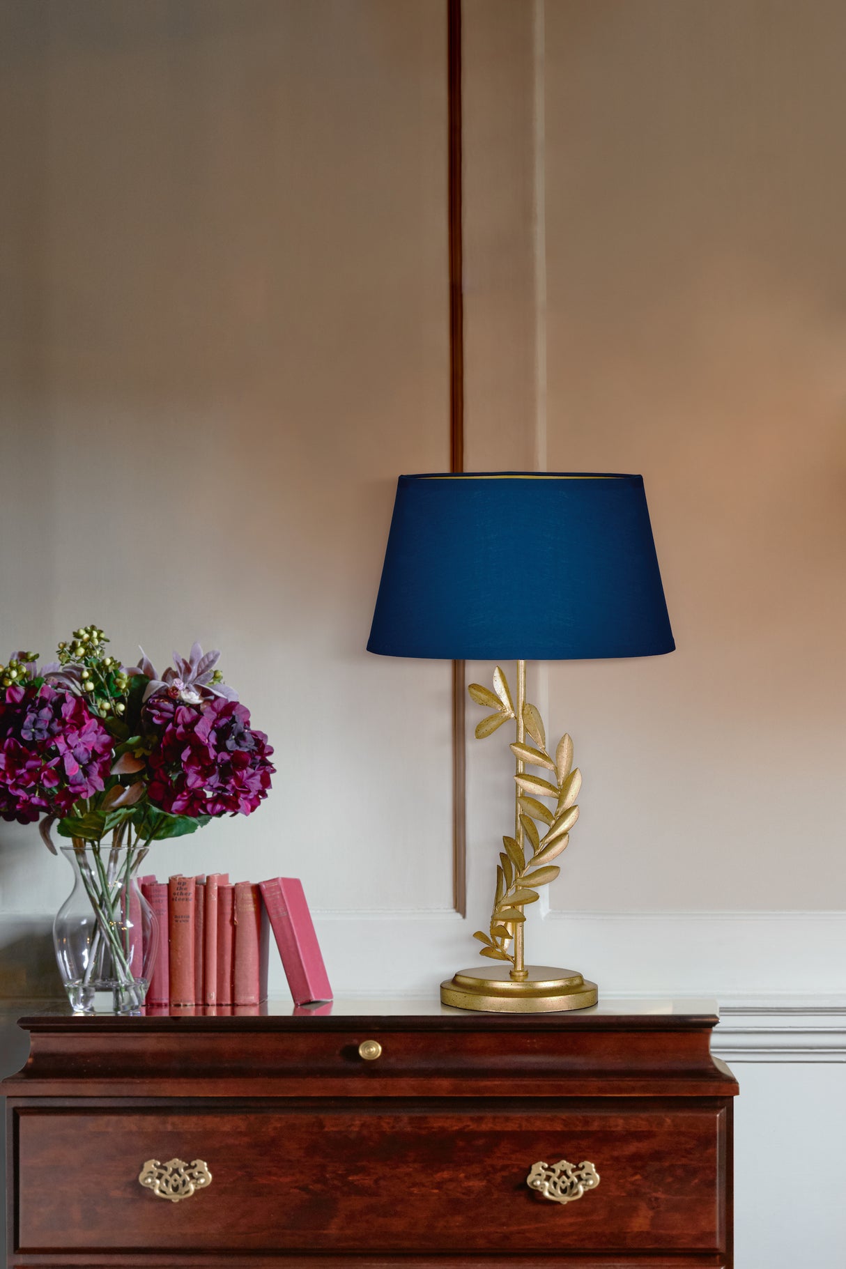 Elegant table lamp with blue shade and gold leaf base on wooden chest with pink books and purple flowers