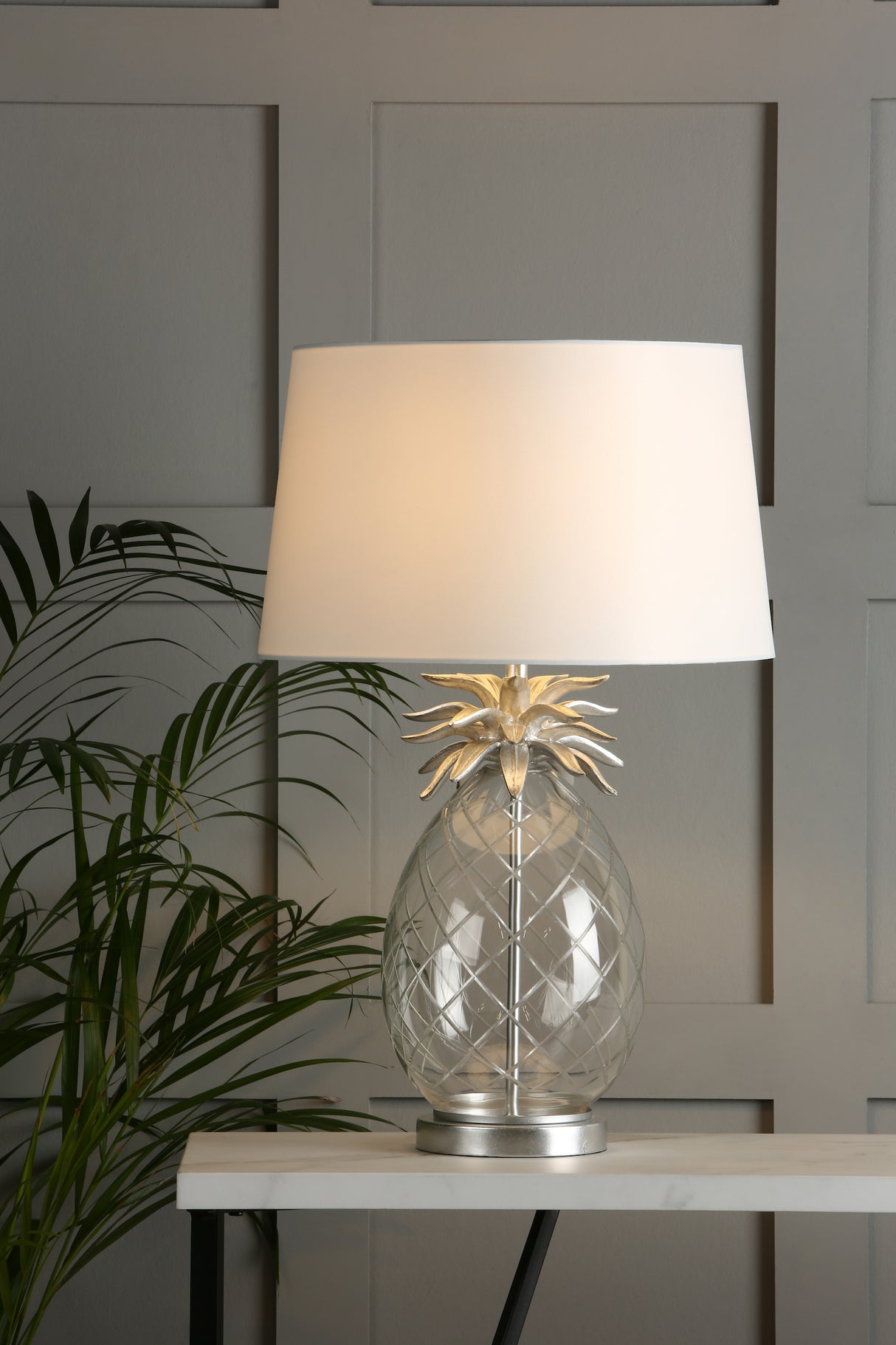 Decorative glass pineapple table lamp with white shade on marble table against paneled wall