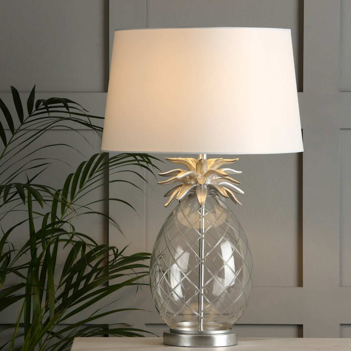 Glass pineapple table lamp with silver leaves and white lampshade on wooden surface