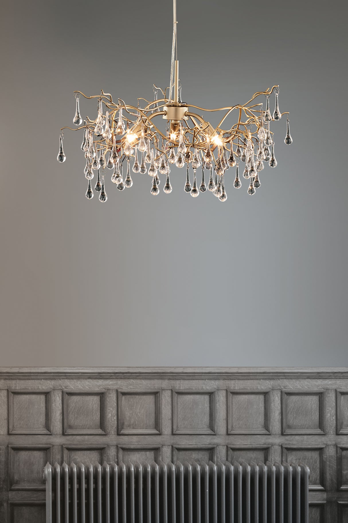 Modern chandelier with gold branches and glass droplets above gray wainscoted wall and radiator