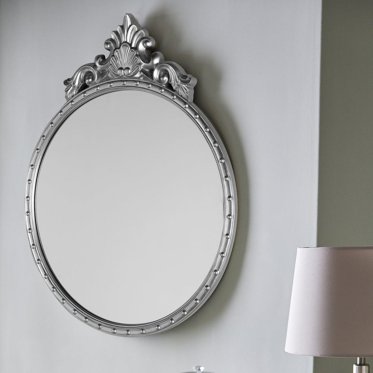 Decorative round silver wall mirror with ornate top detail next to white table lamp on gray wall