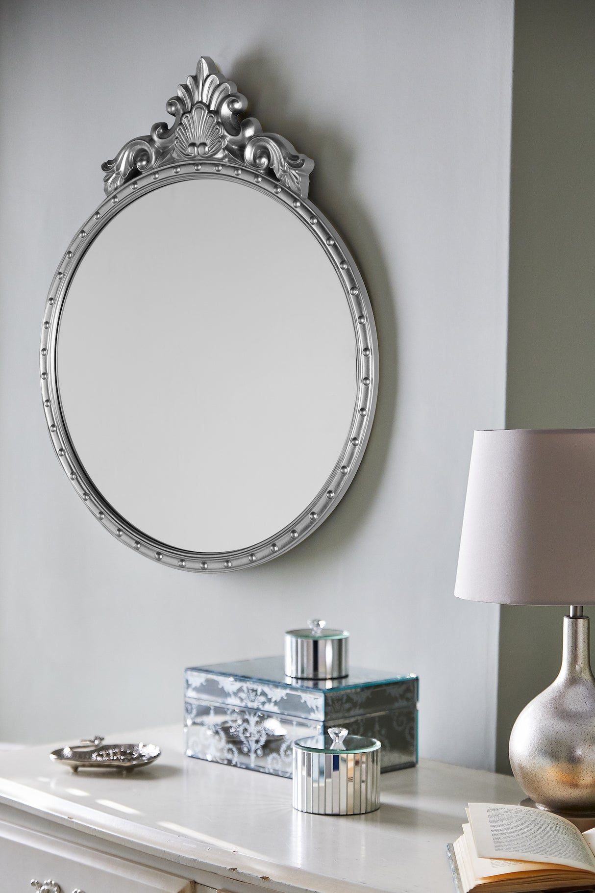 Ornate silver round wall mirror above white dresser with silver containers, lamp, and open book